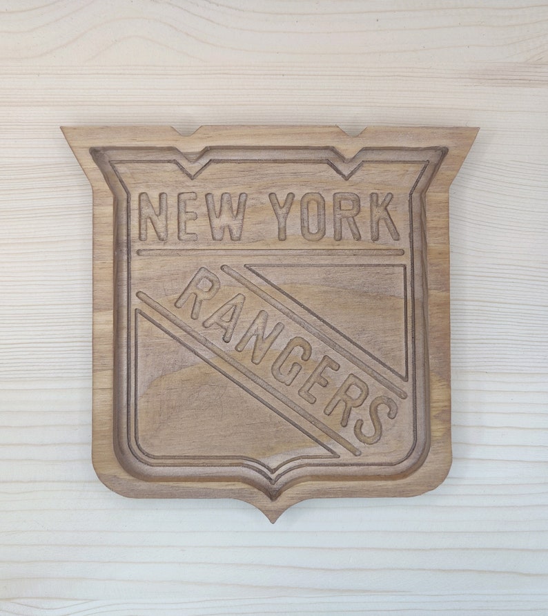 Puede incluir: Un letrero de madera con el logotipo de los New York Rangers. El logotipo est&aacute; tallado en la madera e incluye el nombre del equipo y una franja diagonal.