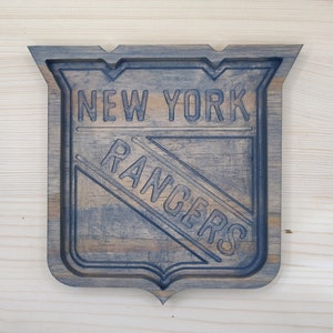 Puede incluir: Un letrero de madera con un logotipo azul y blanco de los New York Rangers. El letrero tiene forma de escudo y un acabado de madera desgastado.