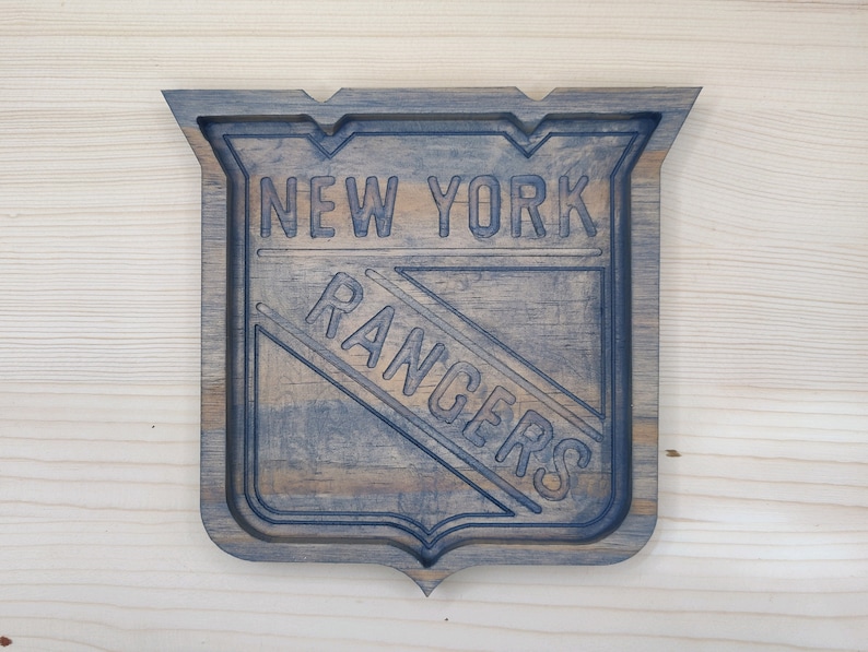 Puede incluir: Un letrero de madera con un logotipo azul y blanco de los New York Rangers. El letrero tiene forma de escudo y un acabado de madera desgastado.