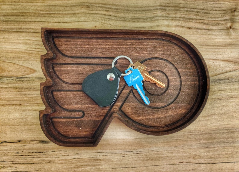 May include: A dark brown wooden key holder shaped like a hockey team's logo. The holder has a black leather key fob with a silver button and a set of keys with a blue tag that says "Home".