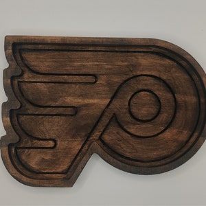 May include: A wooden wall decor in the shape of the Philadelphia Flyers hockey team logo. The logo is brown and features a stylized letter "P" with a wing design.