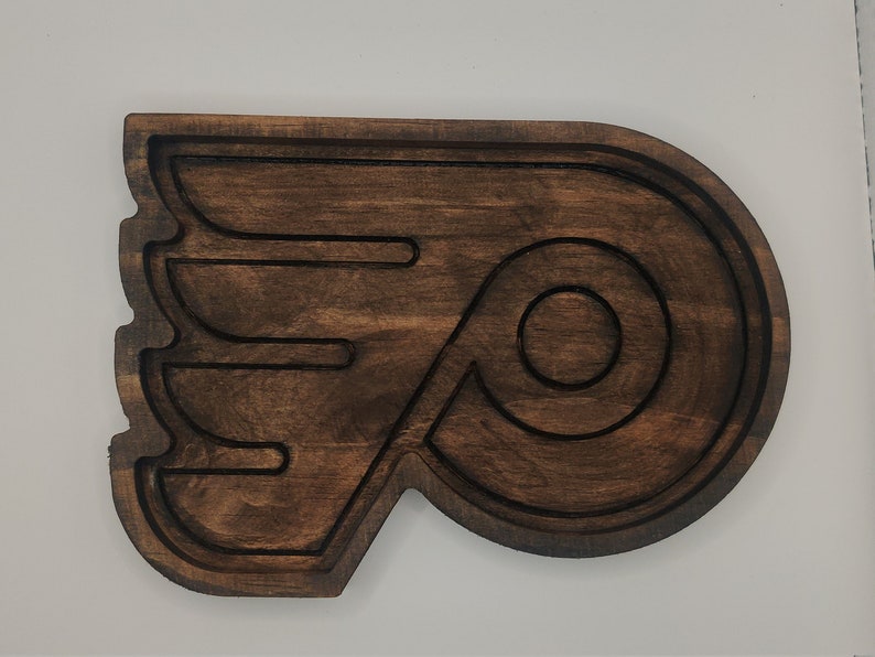 May include: A wooden wall decor in the shape of the Philadelphia Flyers hockey team logo. The logo is brown and features a stylized letter "P" with a wing design.