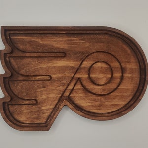 May include: A wooden wall decor in the shape of the Philadelphia Flyers logo. The logo is brown and features a stylized wing with a circle inside.