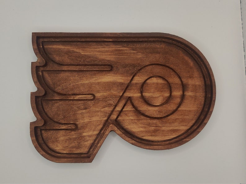 May include: A wooden wall decor in the shape of the Philadelphia Flyers logo. The logo is brown and features a stylized wing with a circle inside.