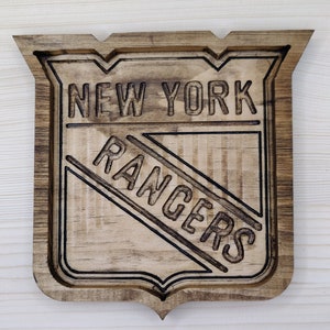 Puede incluir: Un letrero de madera con el logotipo de los New York Rangers. El logotipo es un escudo con las palabras "New York Rangers" en su interior. El letrero es marr&oacute;n y tiene un aspecto desgastado.