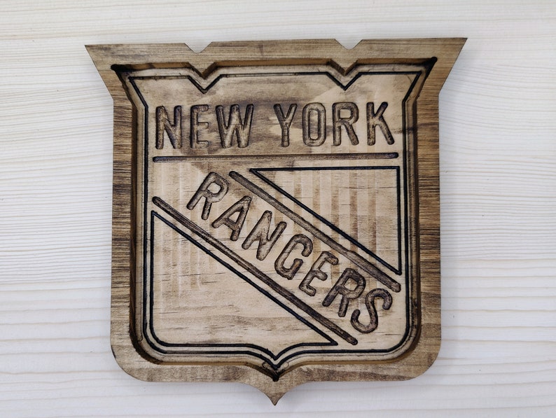 Puede incluir: Un letrero de madera con el logotipo de los New York Rangers. El logotipo es un escudo con las palabras "New York Rangers" en su interior. El letrero es marr&oacute;n y tiene un aspecto desgastado.