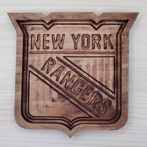 Puede incluir: Un letrero de madera con el logotipo de los New York Rangers. El logotipo es un escudo con las palabras "New York Rangers" en color marr&oacute;n oscuro.