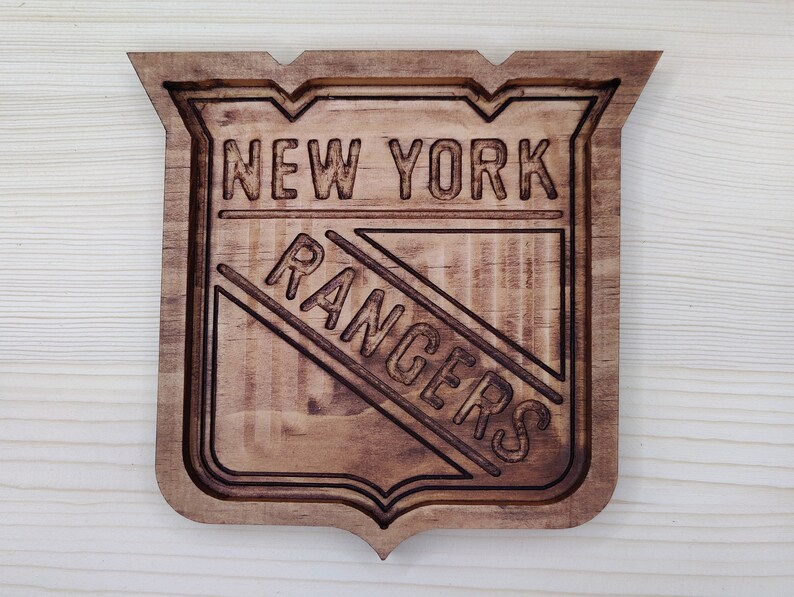 Puede incluir: Un letrero de madera con el logotipo de los New York Rangers. El logotipo es un escudo con las palabras "New York Rangers" en color marr&oacute;n oscuro.