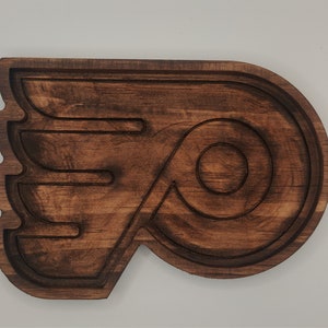 May include: A wooden carving of the Philadelphia Flyers logo. The logo is a stylized letter "P" with a wing on the left side. The carving is dark brown and has a smooth finish.