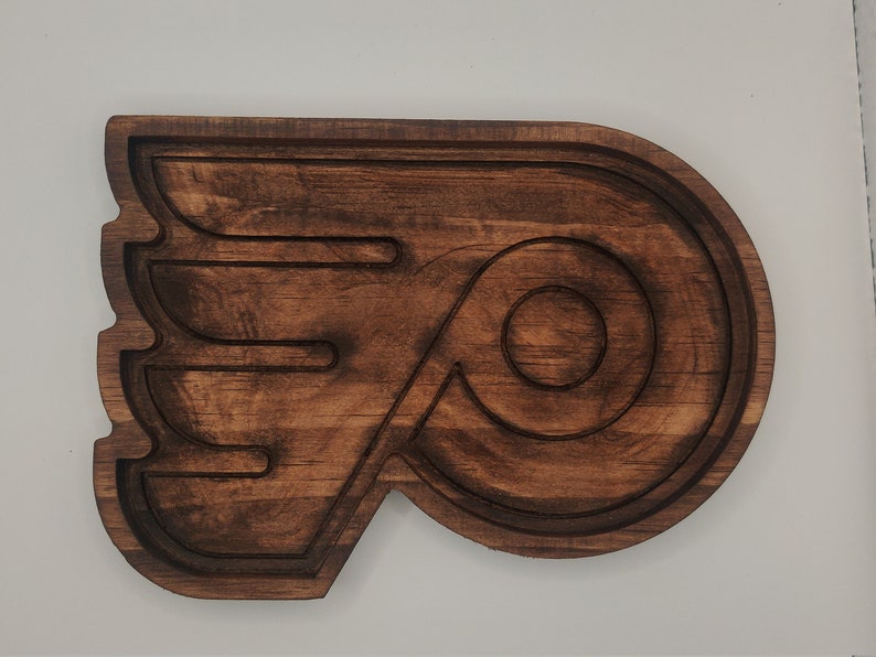 May include: A wooden carving of the Philadelphia Flyers logo. The logo is a stylized letter "P" with a wing on the left side. The carving is dark brown and has a smooth finish.