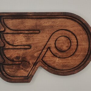 May include: A wooden wall decor in the shape of the Philadelphia Flyers logo. The logo is brown and features a stylized wing with a circular 'P' inside.