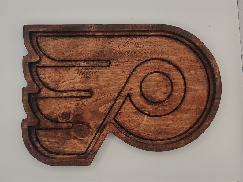 May include: A wooden wall decor in the shape of the Philadelphia Flyers logo. The logo is brown and features a stylized wing with a circular 'P' inside.