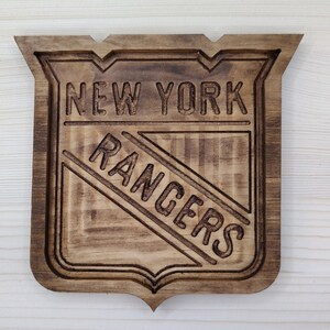 Puede incluir: Un letrero de madera con el logotipo de los New York Rangers. El logotipo est&aacute; tallado en la madera y presenta el nombre del equipo y un palo de hockey estilizado.