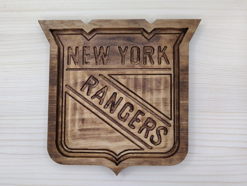 Puede incluir: Un letrero de madera con el logotipo de los New York Rangers. El logotipo est&aacute; tallado en la madera y presenta el nombre del equipo y un palo de hockey estilizado.