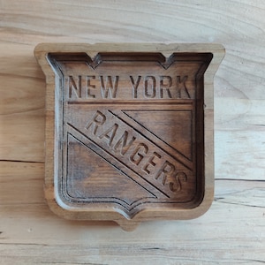 Puede incluir: Una bandeja de madera con un logotipo de los New York Rangers tallado. El logotipo tiene forma de escudo con las palabras "New York" sobre la franja diagonal y "Rangers" debajo de la franja.