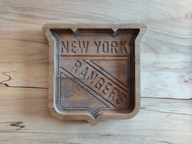 Puede incluir: Una bandeja de madera con un logotipo de los New York Rangers tallado. El logotipo tiene forma de escudo con las palabras "New York" sobre la franja diagonal y "Rangers" debajo de la franja.
