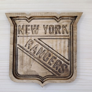 Puede incluir: Un letrero de madera con el logotipo de los New York Rangers. El logotipo es un escudo con las palabras "New York" en la parte superior y "Rangers" en diagonal a trav&eacute;s del escudo.