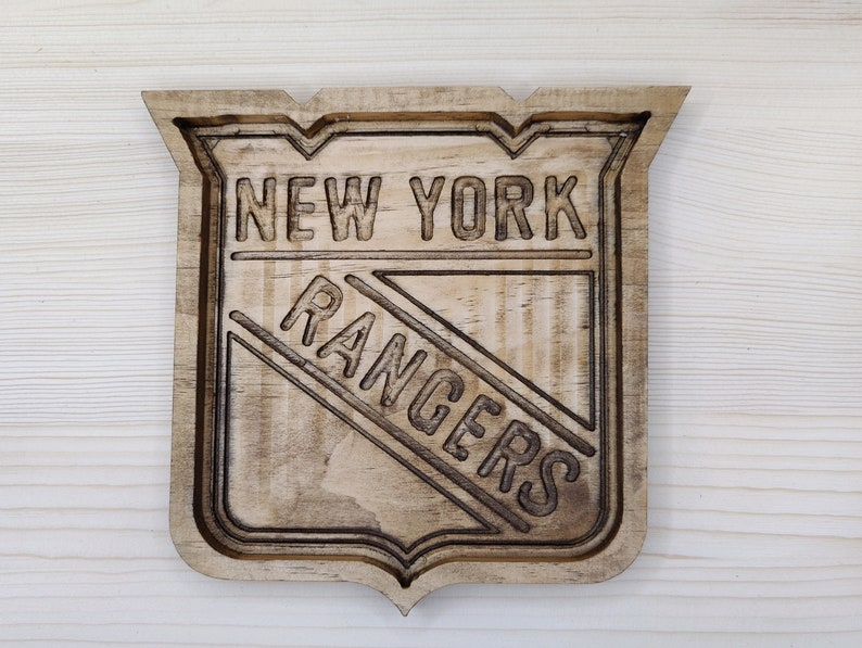 Puede incluir: Un letrero de madera con el logotipo de los New York Rangers. El logotipo es un escudo con las palabras "New York" en la parte superior y "Rangers" en diagonal a trav&eacute;s del escudo.