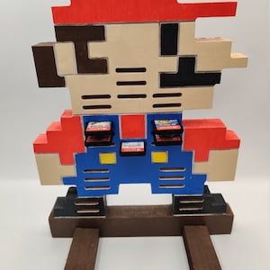 May include: A wooden Nintendo Switch game cartridge holder shaped like a pixelated Super Mario character. The holder is painted red, brown, beige, blue, and black. The holder has slots for 10 game cartridges.