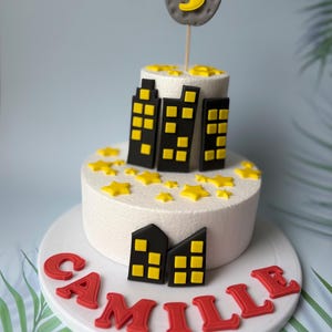 Puede incluir: Un pastel blanco de dos pisos decorado con paisajes urbanos de fondant negro y amarillo, estrellas amarillas y una luna de fondant gris con un número 5 amarillo. El pastel está sobre un soporte para pastel blanco con el nombre "Camille" escrito en letras de fondant rojo.