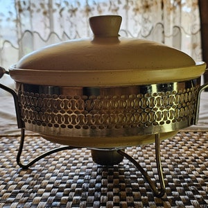 Puede incluir: Un plato de servir de cerámica amarillo con una base de metal dorado y un patrón decorativo. El plato tiene una tapa y dos asas.