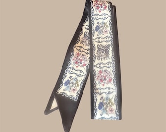 May include: A black scarf with a floral pattern in red, blue, and white. The scarf has a cream-colored border with a paisley design and a black background. The scarf is made of a soft, silky material.