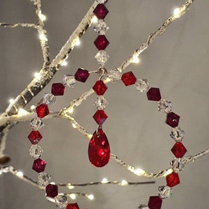 May include: A circular ornament constructed from alternating clear and deep red faceted beads. A vertical strand of beads hangs from the top of the circle, terminating in a large, teardrop-shaped red crystal pendant suspended in the centre.