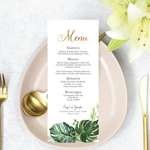 May include: A wedding menu with a tropical theme. The menu is printed on a white card with gold lettering and features a watercolor illustration of green tropical leaves. The menu includes starters, mains, desserts, and beverages. The text on the menu reads: "Menu", "Starters", "Salmon & John Dory Terrine", "Red Onion and Aubergine Tart", "Mains", "Black Angus chopped steak with roasted portabello mushrooms and sautéed onions", "Pan fried lemon Chicken Morsels with créme craiche and lemon zests", "Desserts", "Dark Chocolate Pudding", "Vanilla ice cream", "Glazed Lemon Tart", "Beverages", "Lemonade Iced Tea", "Red Wine", "Tea & Coffee", "Hagal & Joseph", "CLIFTON HILL GOLF CLUB", "APRIL 30, 2015".