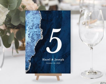 May include: A table number card with a blue and silver design, displaying the number 5 in white. The card includes the names "Hazel & Joseph" and the date "October 08, 20XX". The card is propped up on a small wooden easel, set on a white tablecloth.