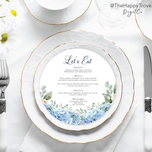 Peut inclure: Une assiette blanche avec un bord doré présente une illustration aquarelle d'hortensias bleus et de feuilles vertes. L'assiette comporte un menu imprimé avec le titre "Let's Eat" et liste les entrées, les plats principaux, les desserts et les boissons.