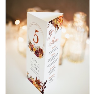 Puede incluir: Una tarjeta de número de mesa con un círculo dorado y el número 5 en cobre. La tarjeta tiene un diseño floral con colores otoñales y hojas. El texto "Hazel y Joseph" está impreso en la tarjeta, junto con la fecha "20 de octubre de 20XX" y la ubicación "Capilla Gold Club". La tarjeta también incluye un menú con entrantes, platos principales, postres y bebidas.