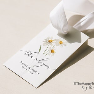 May include: White wedding favor tag with a bouquet of white daisies and green stems. The tag says "Thank you" in cursive script and "Hazel & Joseph 12.04.20XX" in black text. The tag is tied with a white ribbon.