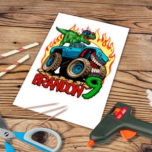 May include: A blue monster truck with flames and a green dinosaur on top. The truck has a toothy grin and the words "BRANDON 9" are written below it.