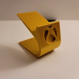 May include: A gold-colored plastic stand for a video game console. The stand has a cutout of the Xbox logo.