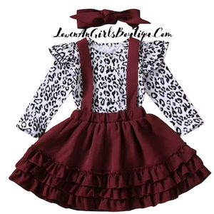 May include: A three-piece outfit for a child, featuring a white and black leopard print long-sleeved shirt, a burgundy skirt with multiple ruffles, and a burgundy headband. The skirt has suspenders.