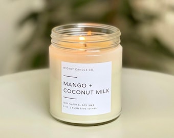May include: A clear glass jar candle with a white label that reads "WICKRY CANDLE CO. MANGO + COCONUT MILK 100% NATURAL SOY WAX 8 OZ | BURN TIME 40 HRS". The candle is lit and the flame is visible.