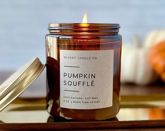 Peut inclure: Une bougie en pot de verre ambré allumée avec un couvercle doré, étiquetée "Pumpkin Soufflé". La bougie est posée sur un plateau doré. L'étiquette indique "100% Natural Soy Wax" et "8 oz | Burn Time 40 hrs."