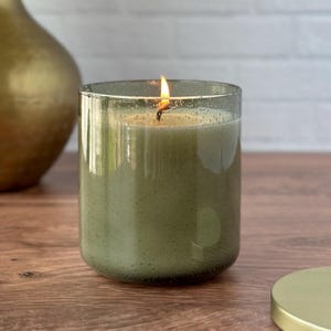 May include: A lit green candle in a clear glass jar with a gold lid. The candle sits on a wooden surface, with a gold vase and dried plants in the background. The candle flame is visible.