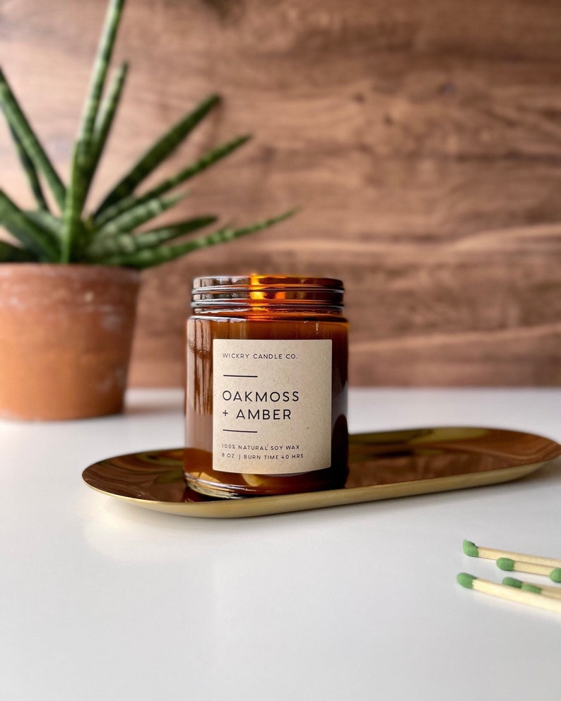 May include: A brown glass candle jar with a label that reads "WICKRY CANDLE CO. OAKMOSS + AMBER 100% NATURAL SOY WAX 8 OZ | BURN TIME 40 HRS" The candle jar is sitting on a gold tray.