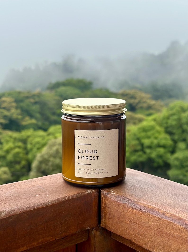 May include: A brown glass candle jar with a gold lid, labeled "Cloud Forest" by Wickry Candle Co. The label indicates the candle is made with 100% natural soy wax and has an 8 oz volume with a 40-hour burn time. The candle sits on a wooden surface.
