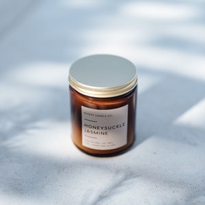 May include: A brown glass candle jar with a gold lid. The label reads "Wickery Candle Co." and "Honeysuckle Jasmine". The candle is made with natural soy wax.
