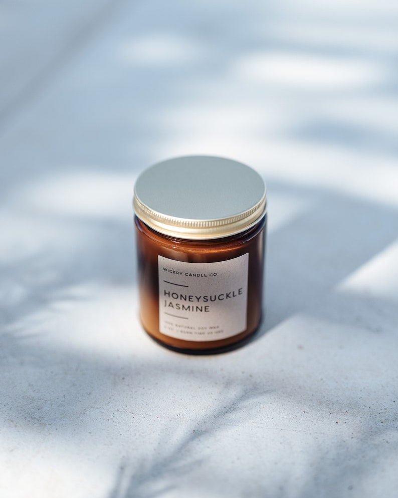 May include: A brown glass candle jar with a gold lid. The label reads "Wickery Candle Co." and "Honeysuckle Jasmine". The candle is made with natural soy wax.