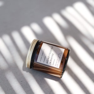 May include: A brown glass candle jar with a gold lid. The label reads "Wickry Candle Co. Honeysuckle Jasmine 100% Natural Soy Wax 8 oz | Burn Time 40 Hrs".