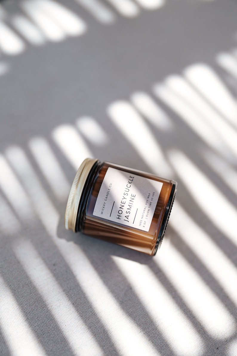 May include: A brown glass candle jar with a gold lid. The label reads "Wickry Candle Co. Honeysuckle Jasmine 100% Natural Soy Wax 8 oz | Burn Time 40 Hrs".