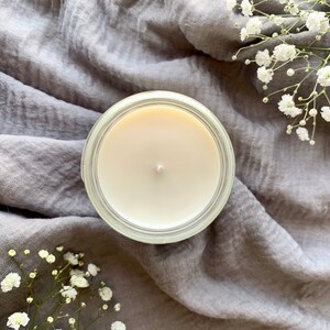 May include: A white candle in a clear glass jar sits on a gray fabric background. The candle is unlit and has a single wick.