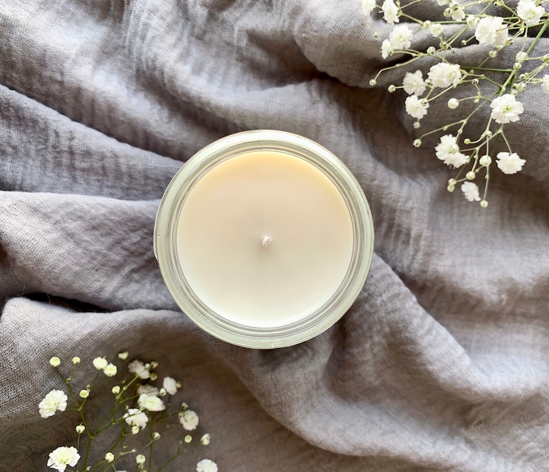May include: A white candle in a clear glass jar sits on a gray fabric background. The candle is unlit and has a single wick.