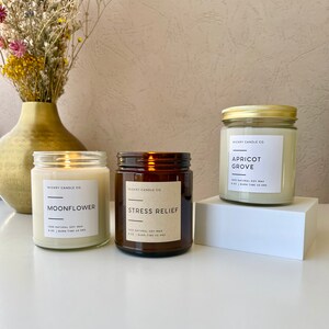 May include: Three amber glass candle jars with white labels. The labels have the text "Wickry Candle Co." and the names of the scents: "Moonflower", "Stress Relief", and "Apricot Grove". Each label also includes the text "100% Natural Soy Wax" and the burn time of the candle.