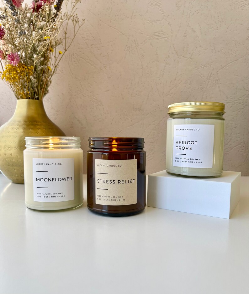 May include: Three amber glass candle jars with white labels. The labels have the text "Wickry Candle Co." and the names of the scents: "Moonflower", "Stress Relief", and "Apricot Grove". Each label also includes the text "100% Natural Soy Wax" and the burn time of the candle.