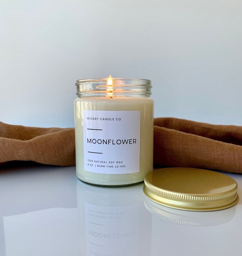 May include: A glass candle jar with a gold lid. The candle is lit and the jar has a white label that reads "WICKRY CANDLE CO." and "MOONFLOWER". The label also states "100% NATURAL SOY WAX" and "8 OZ | BURN TIME 40 HRS".
