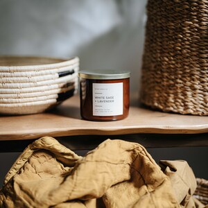 May include: A brown glass candle with a gold lid and a white label that reads "White Sage + Lavender". The candle is sitting on a wooden surface with a woven basket and a yellow blanket.